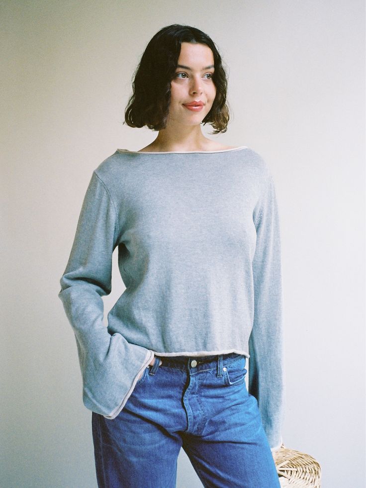 a woman standing in front of a white wall wearing blue jeans and a gray sweater