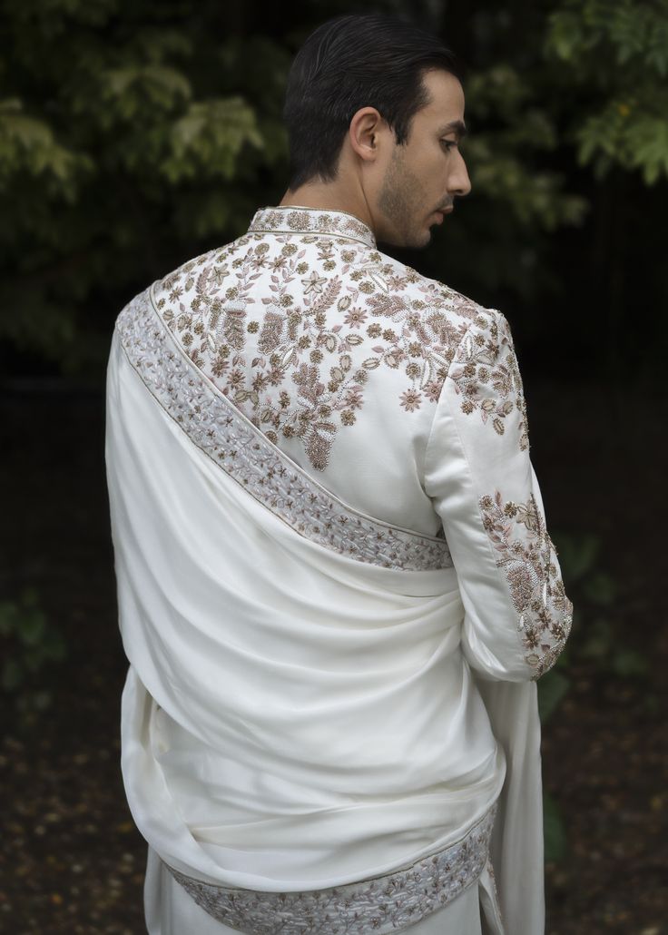 a man wearing a white suit with gold embroidery on the chest and back, standing in front of trees