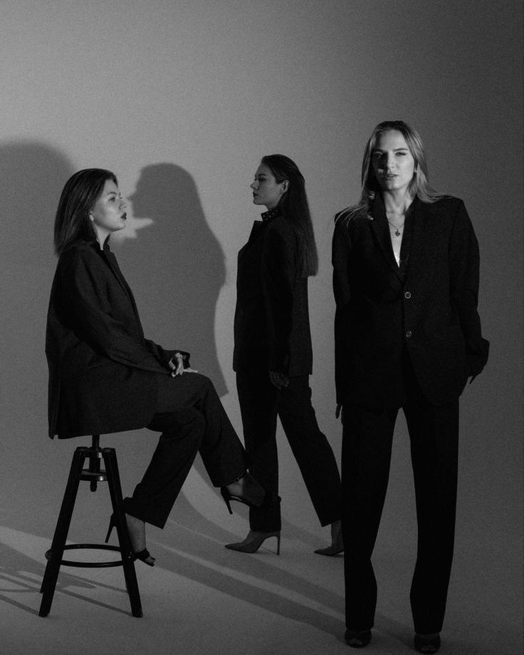 three women standing in front of a white wall and one woman sitting on a stool