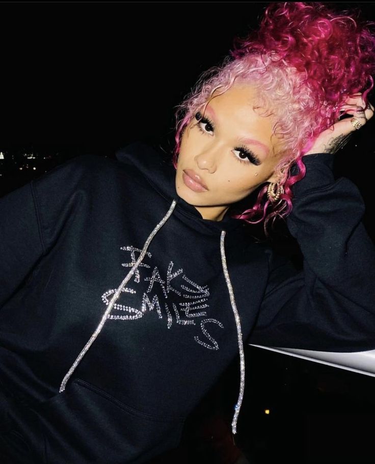 a woman with pink hair wearing a black hoodie and posing for the camera at night