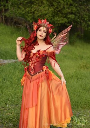 a woman dressed in an orange and red fairy costume standing on grass with her hands behind her back