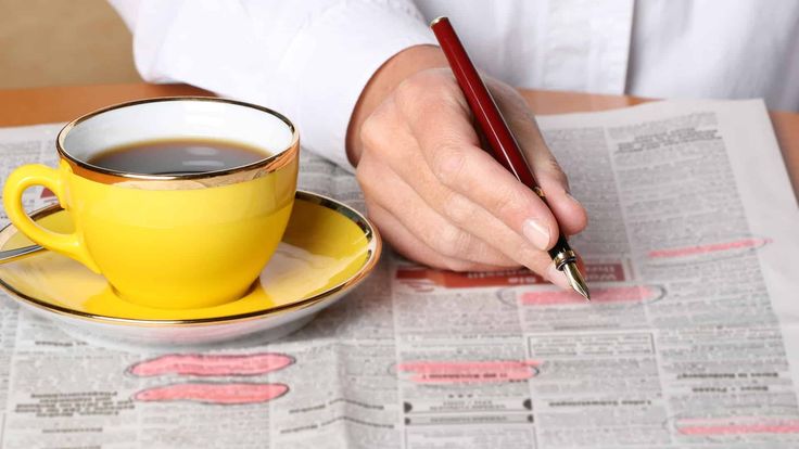 a person writing on a newspaper with a cup of coffee