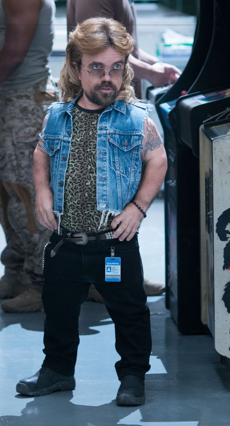 a man with long hair and beard standing in front of a black suitcase wearing a denim vest