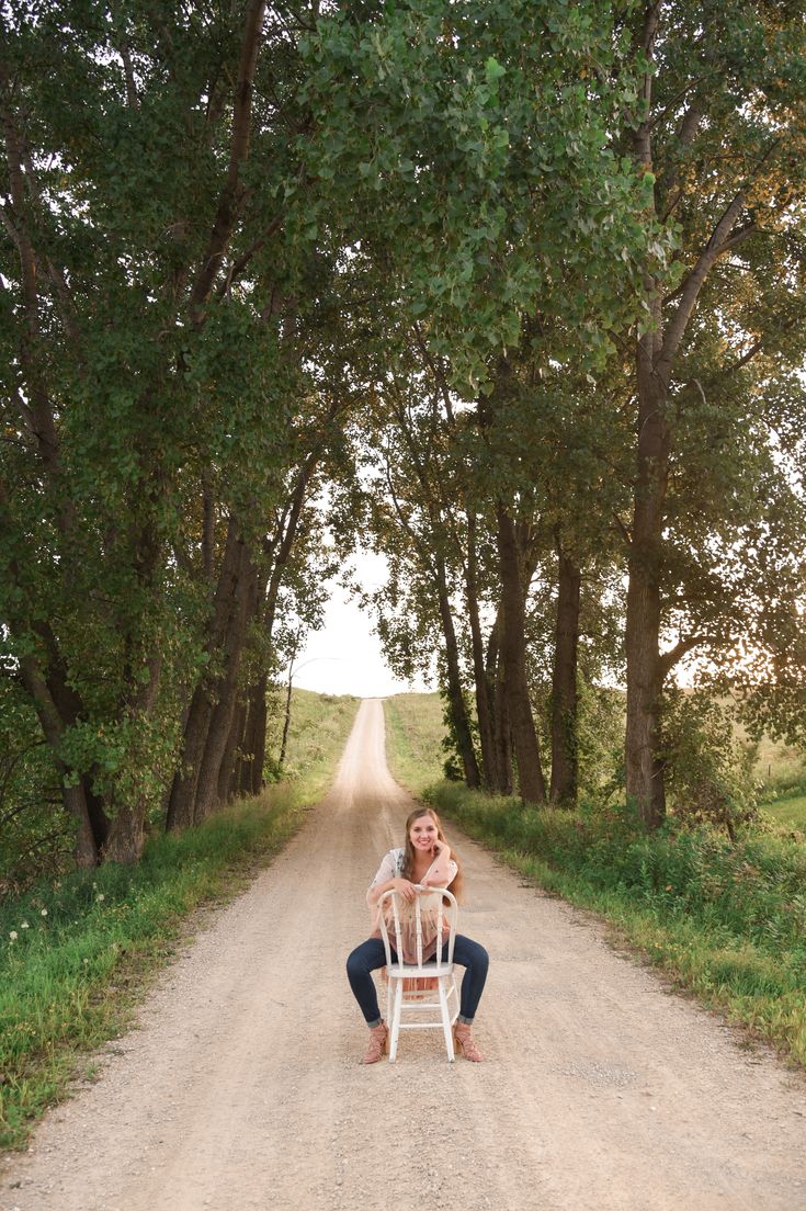 a woman sitting in a chair on the side of a dirt road next to trees