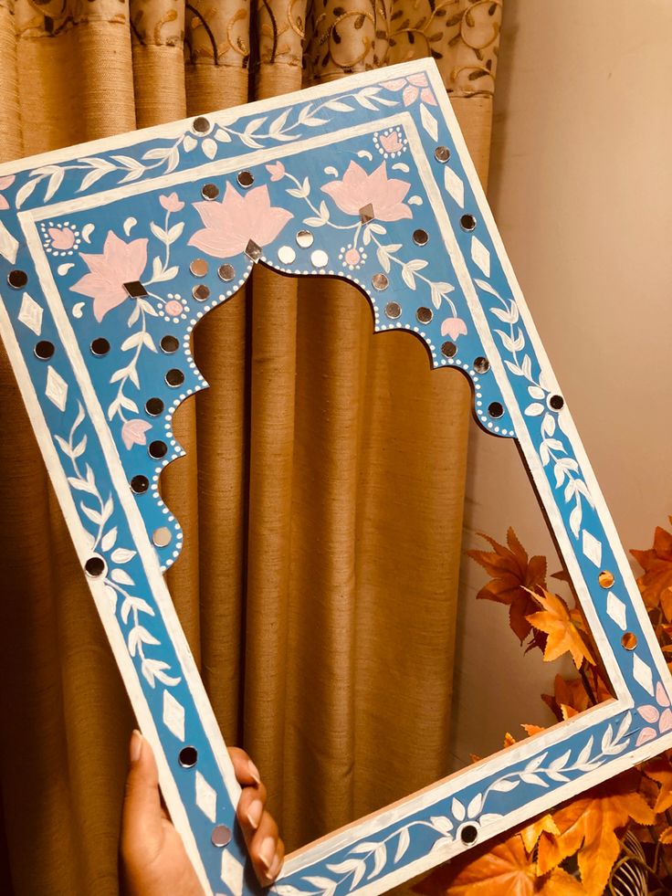 a person holding up a blue and white frame with pink flowers on the edges, in front of a window