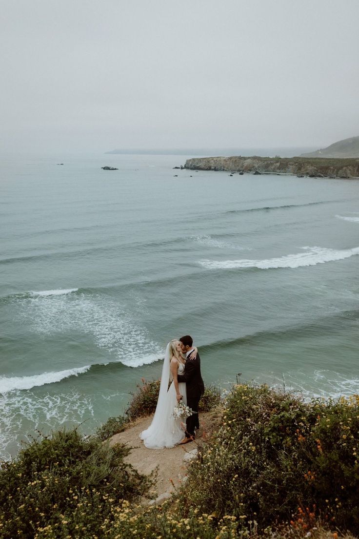 a bride and groom standing on the edge of a cliff overlooking the ocean