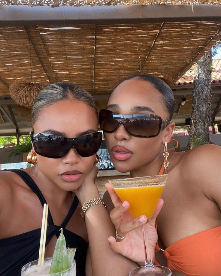 two women pose for the camera with drinks in front of their faces and straw umbrellas behind them