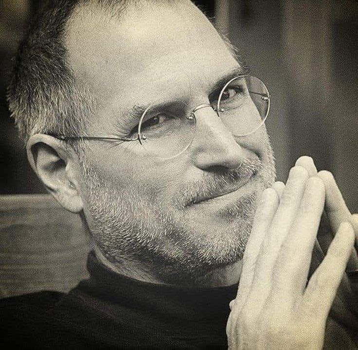 a man with glasses and a goatee looks into the distance while holding his hands to his chin