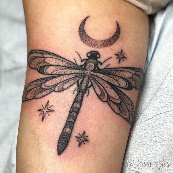 a dragonfly tattoo on the arm with stars and a crescent in the sky above it