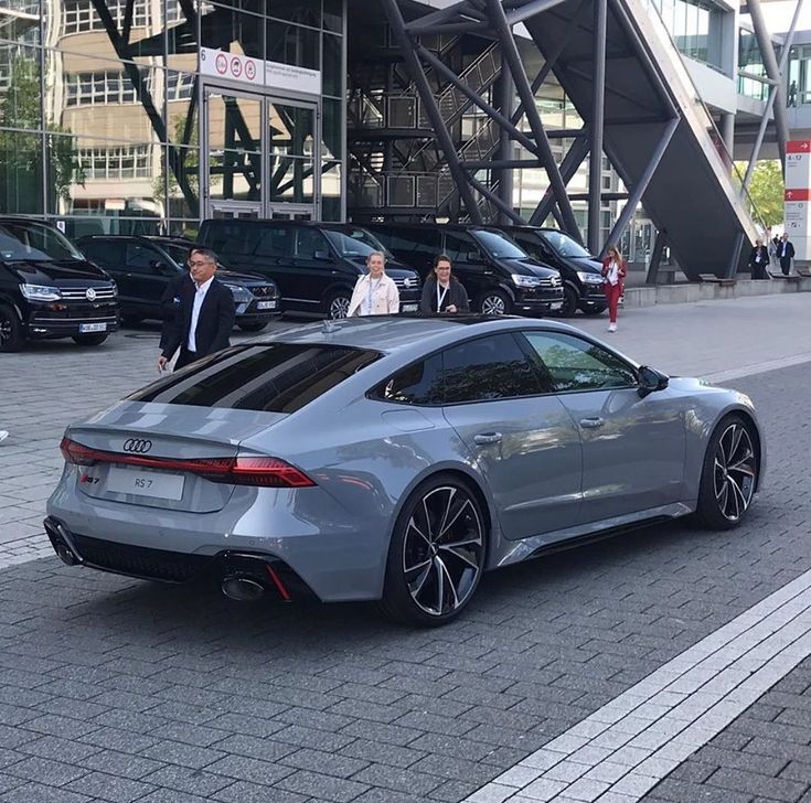 a silver sports car parked in front of a tall building with people walking around it