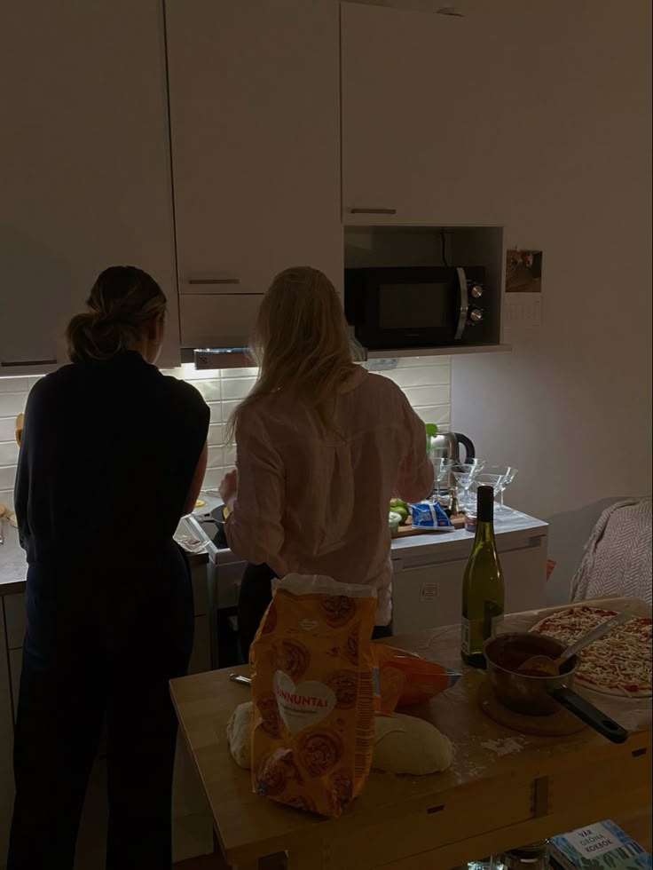 two women standing in a kitchen preparing food and drinking wine on the counter top next to an oven