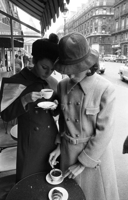 two women standing next to each other while holding coffee cups and looking at their cell phones