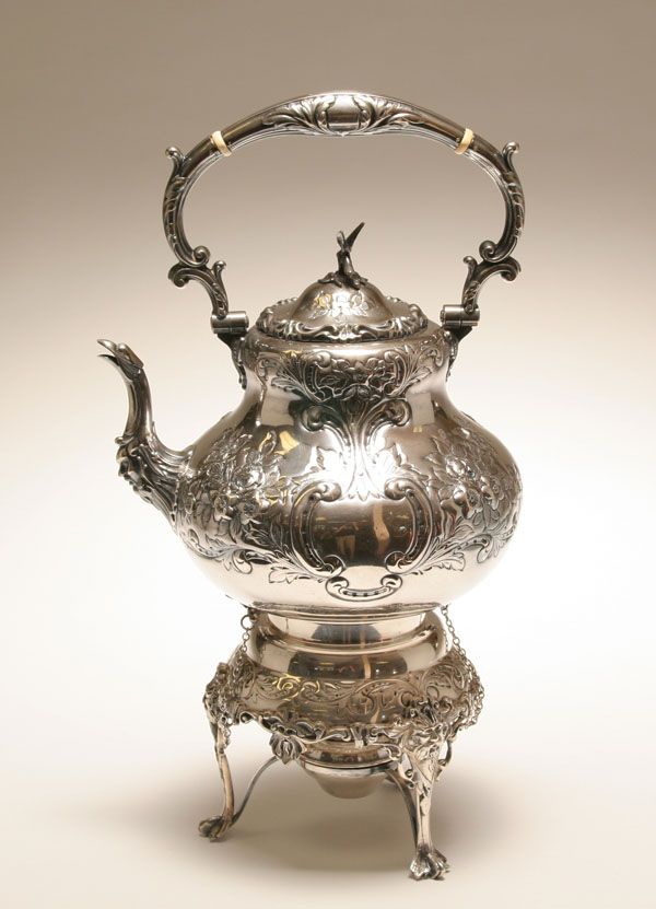 an ornate silver tea pot with handles on a mirrored surface, in front of a white background