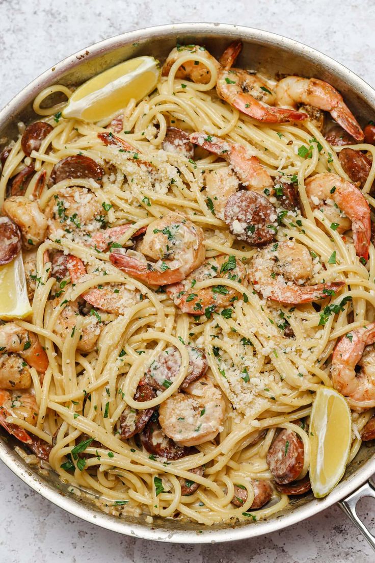 a pan filled with pasta and shrimp on top of a white countertop next to lemon wedges