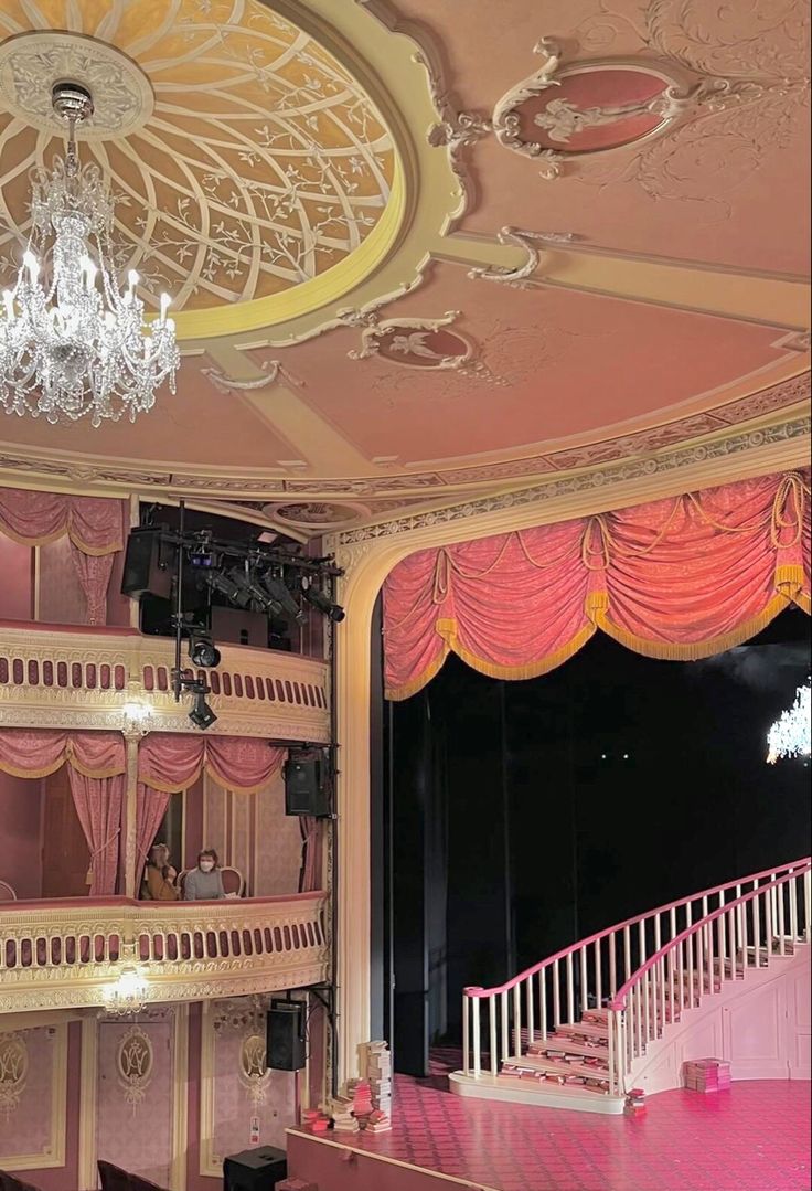 an empty stage with chandelier and pink curtains on the walls, in front of a balcony