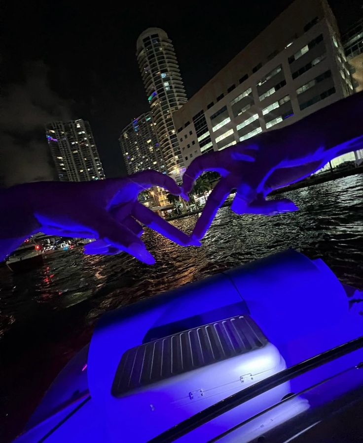 two hands making a heart shape on the back of a boat at night with buildings in the background