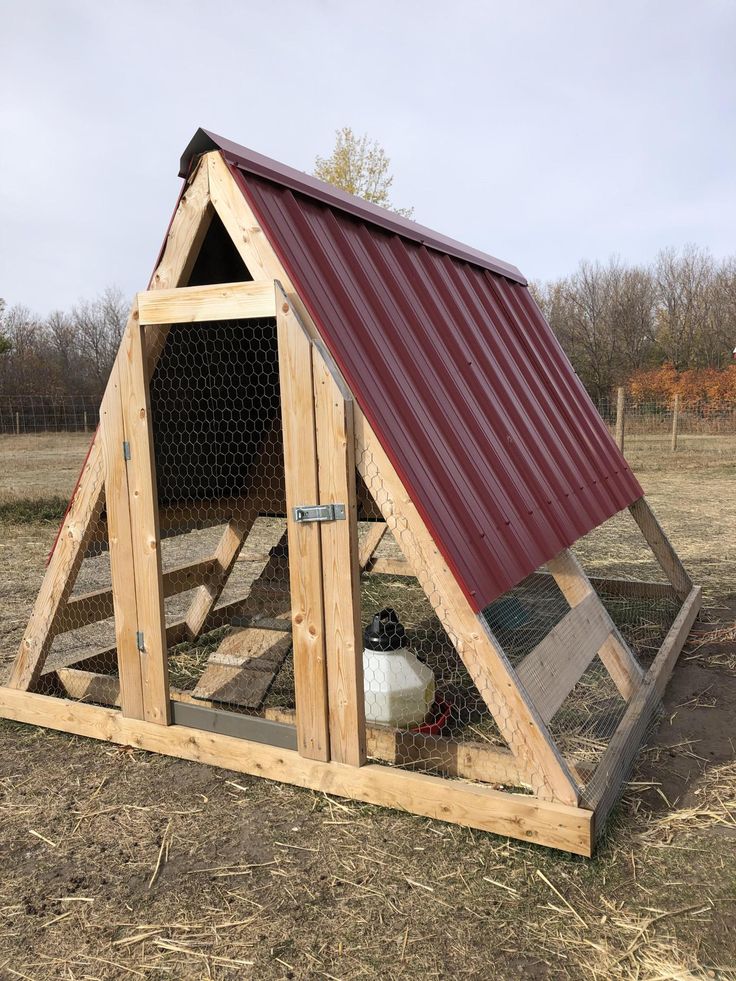 a chicken coop with a red roof and two chickens in the back ground, on top of dry grass