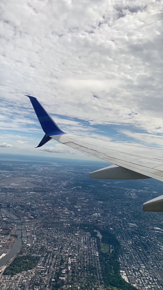 the wing of an airplane flying over a city under a blue sky with white clouds