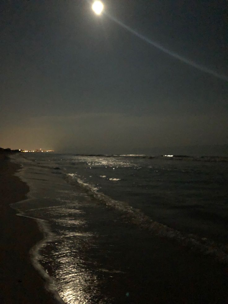 the moon is shining over the water at night on the beach with waves coming in