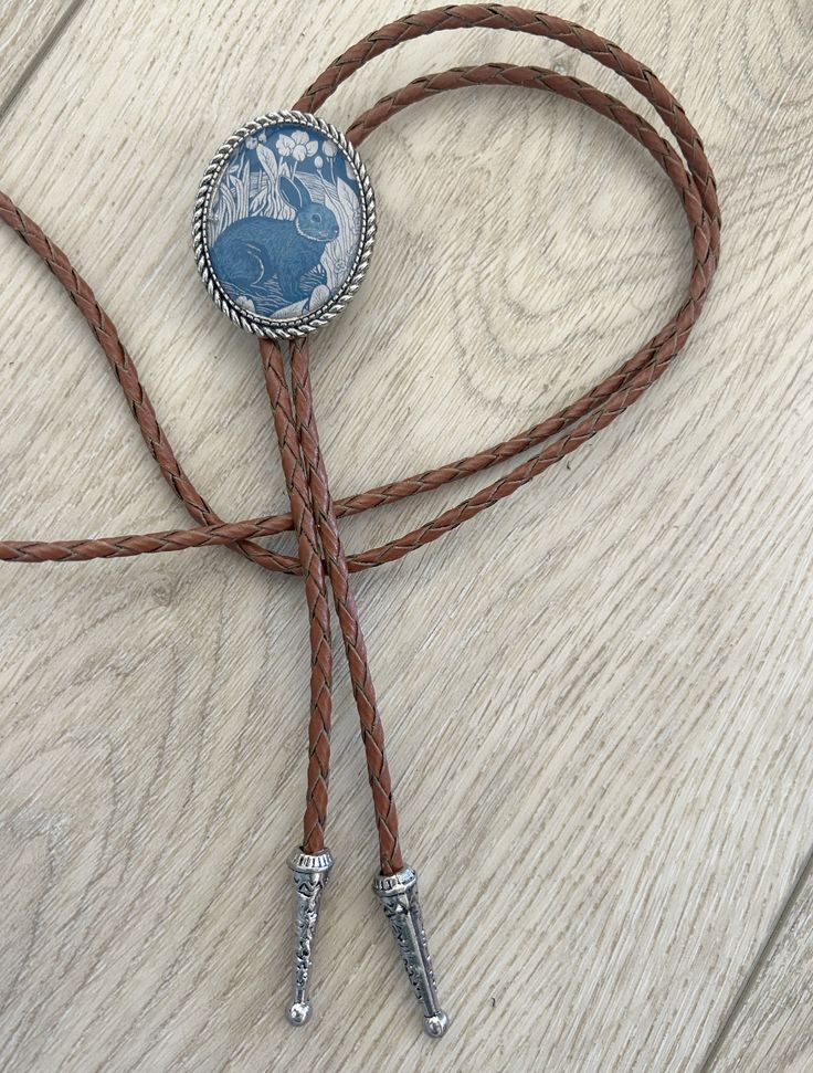 This awesome bolo tie has a vintage rabbit print with a clear resin coating and metal backing. Handmade in our shop!The cord is brown and made of genuine woven leather.Our bolos pair nicely with many of our belt buckles! They make wonderful gifts.The western bolo tie rope length is 39'' ; pendant size is 1 1/2'' x 1 1/4'' Leather Bolo Tie, Vintage Adjustable Hand Tooled Bolo Tie, Adjustable Vintage Hand Tooled Bolo Tie, Vintage Brown Jewelry For Rodeo, Vintage Brown Bolo Tie With Adjustable Length, Handmade Adjustable Bolo Tie For Rodeo, Handmade Adjustable Bolo Ties For Rodeo, Handmade Adjustable Western Bolo Ties, Handmade Adjustable Bolo Ties For Western-themed Events