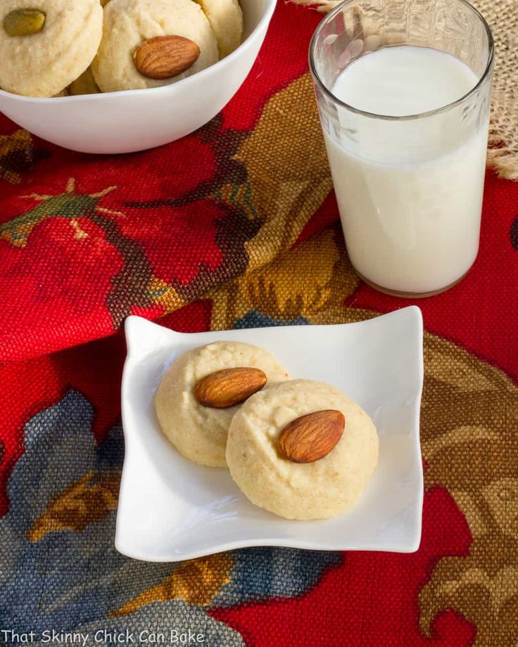 two almond cookies on a white plate next to a glass of milk and bowl of almonds