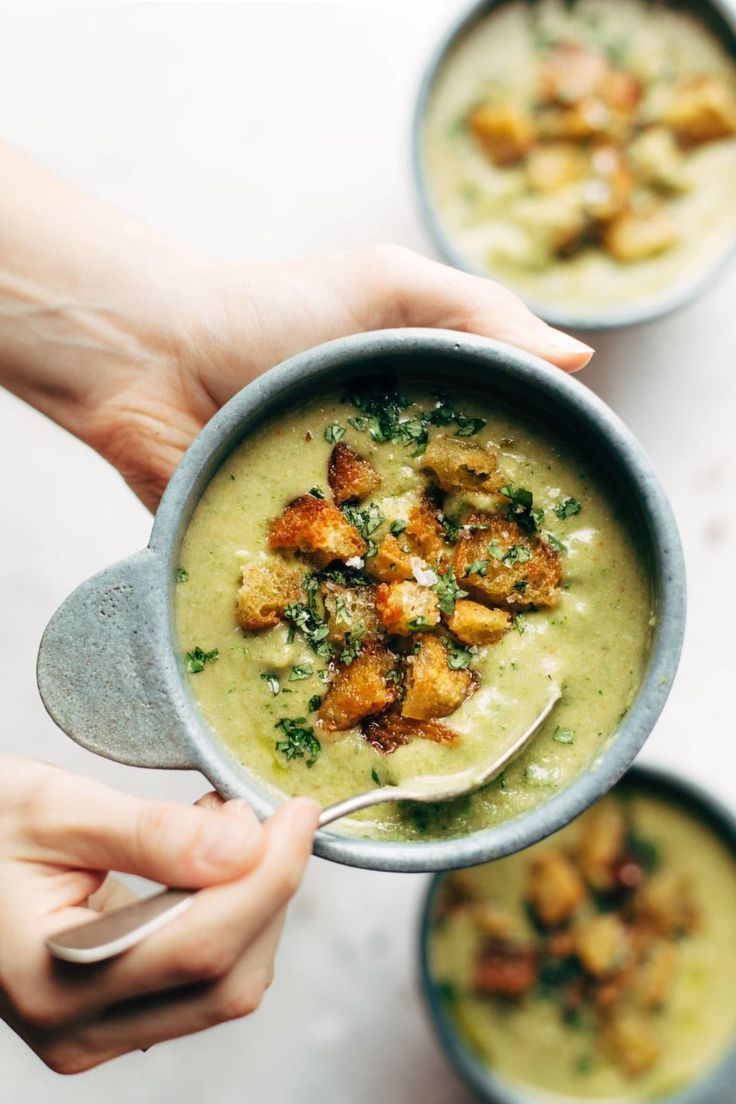 two bowls of soup with broccoli and croutons in them being held by someone's hands