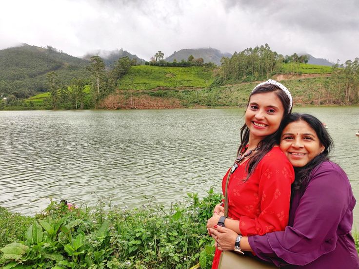 two women standing next to each other in front of a body of water with hills and trees behind them