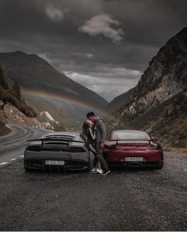 a man and woman kissing next to their cars on the side of the road with a rainbow in the background