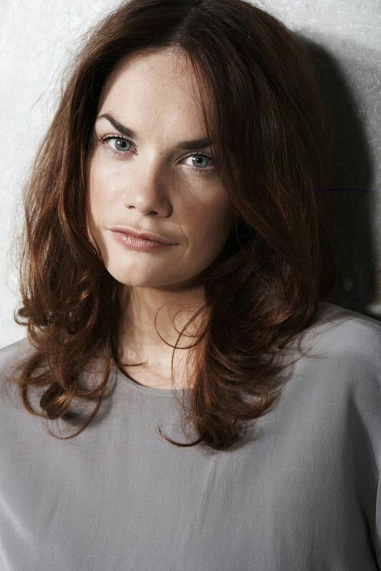 a woman with brown hair and blue eyes is leaning against a wall looking at the camera