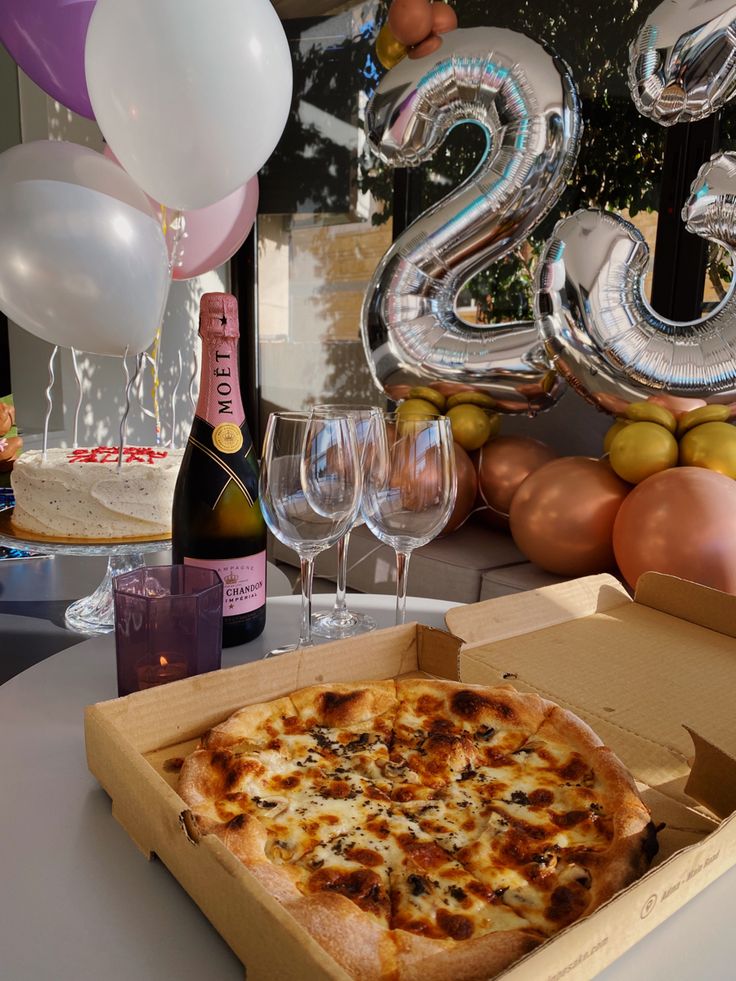 a pizza in a box on a table with balloons and wine glasses next to it