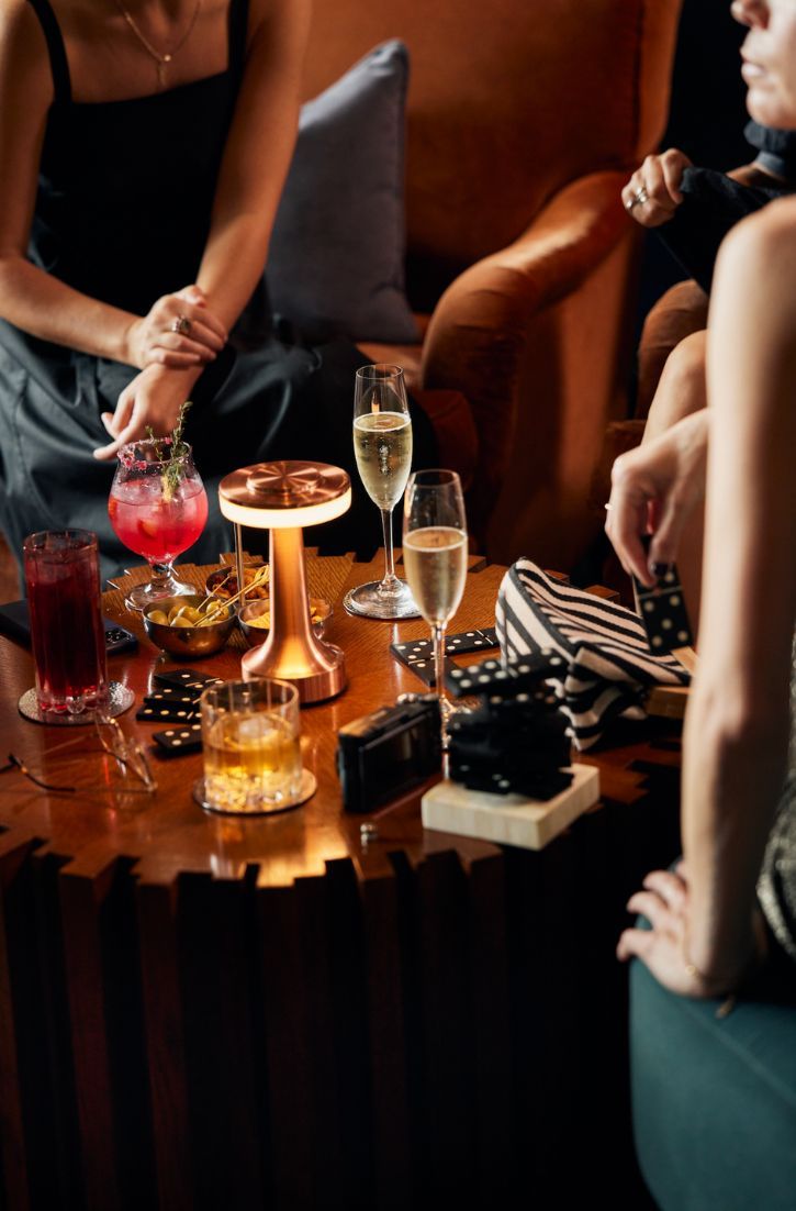 two women sitting at a table with wine glasses and snacks on it, while another woman sits in the background