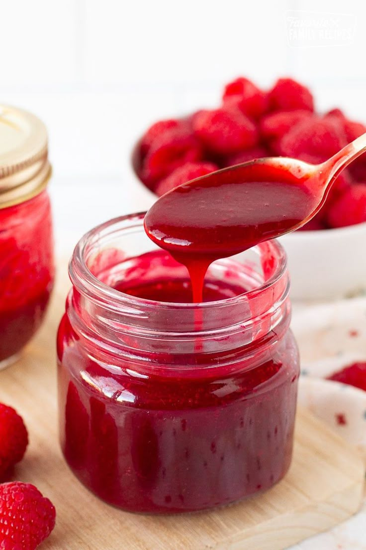 a spoon full of raspberry jam on top of a cutting board with strawberries in the background