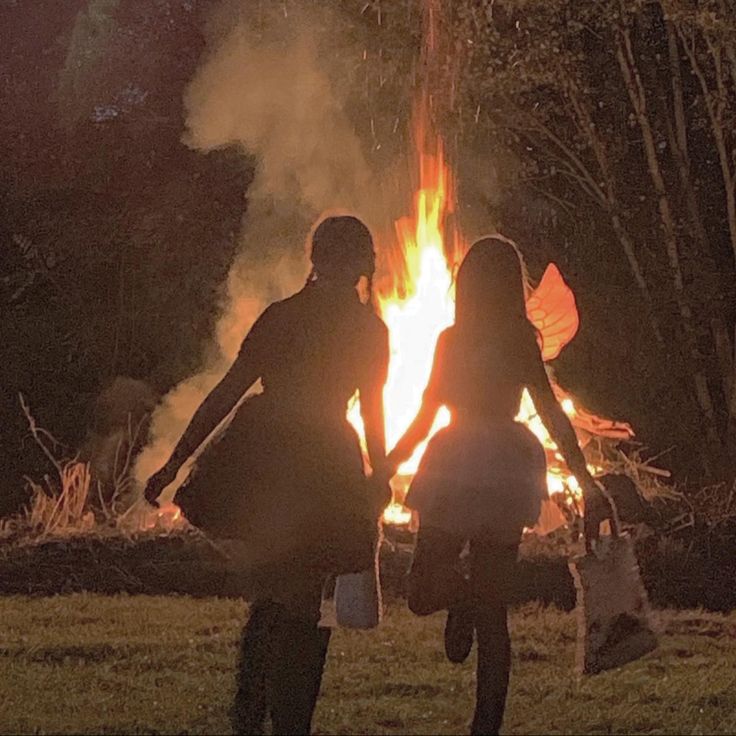 two people standing in front of a fire with their arms around each other and holding hands