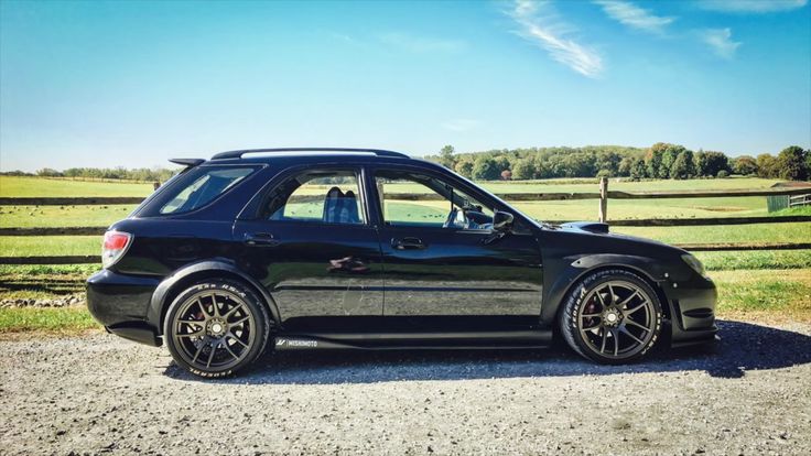 a black car parked in front of a wooden fence on gravel road next to grass and trees