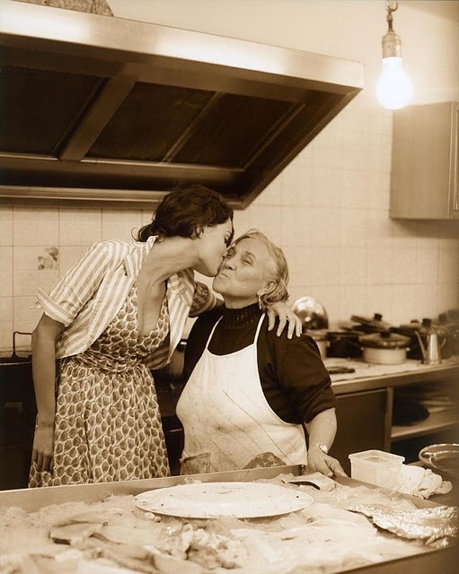 two women are kissing in the kitchen while another woman is preparing food behind them on a counter