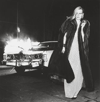 black and white photograph of woman in fur coat next to car on street at night