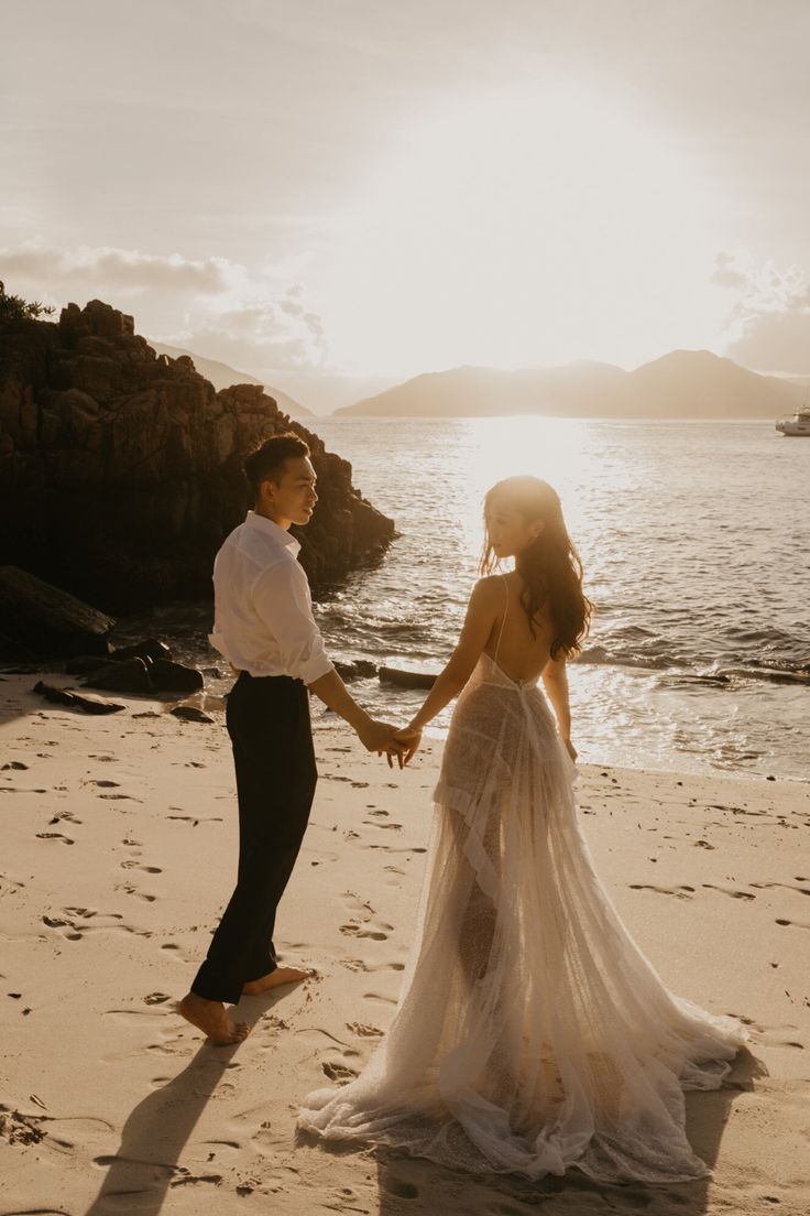 a bride and groom hold hands on the beach at sunset in front of the ocean