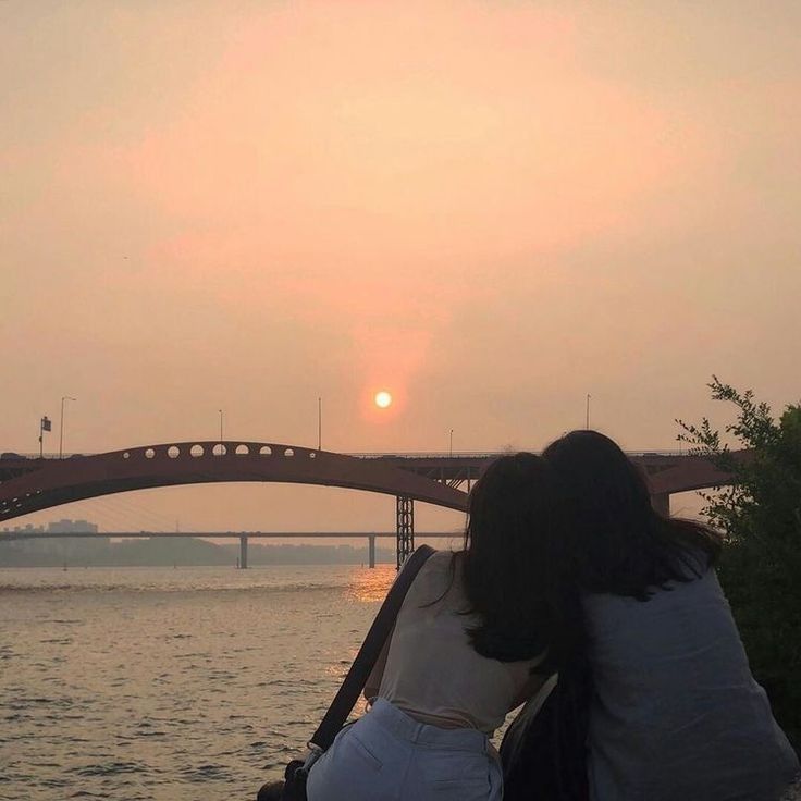 two people sitting on the edge of a bridge watching the sun go down over water