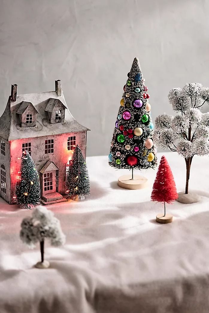 three small christmas trees in front of a house on a table with white snow covered ground