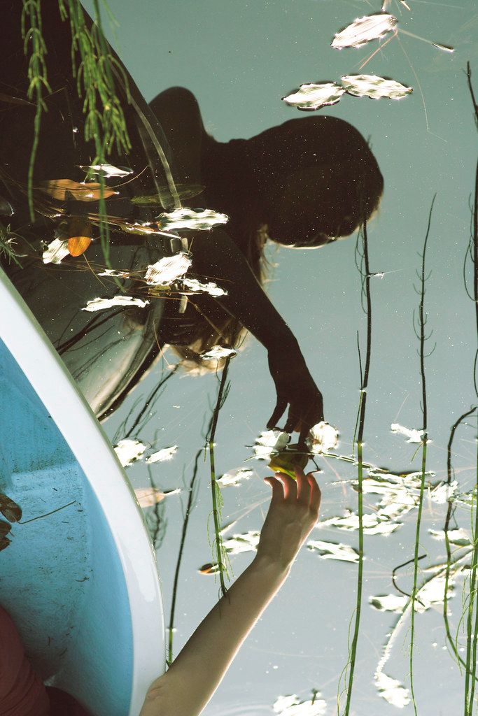 the reflection of a person standing on a boat in water with lily pads and reeds