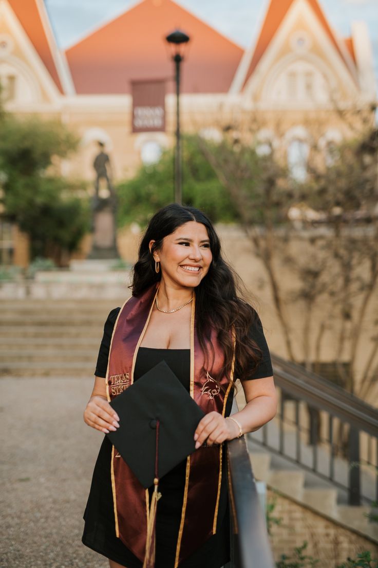 a woman holding a black graduation cap and gown in front of a building with stairs