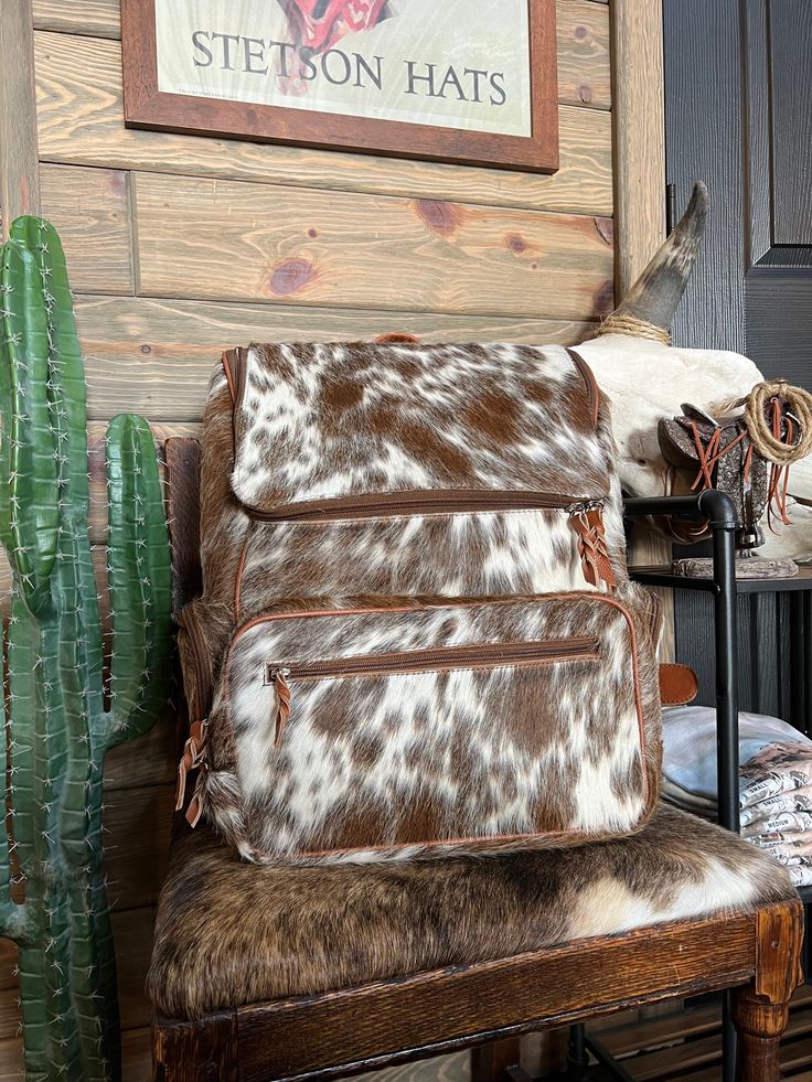 a brown and white cow print backpack sitting on top of a chair next to a cactus