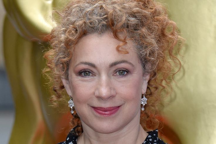 a woman with curly hair and blue eyes smiles at the camera while standing in front of an oscar statue