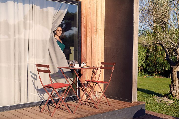 a woman standing in the window of a house with two chairs and a table outside