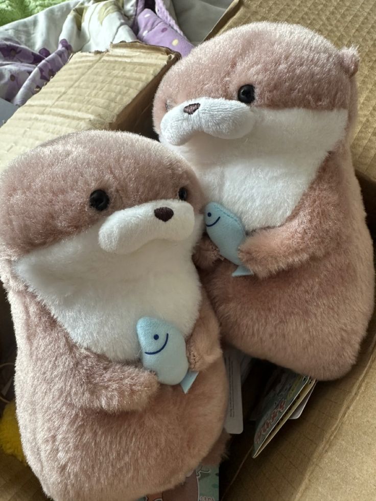 two stuffed animals are sitting in a cardboard box on the floor, one is holding another animal