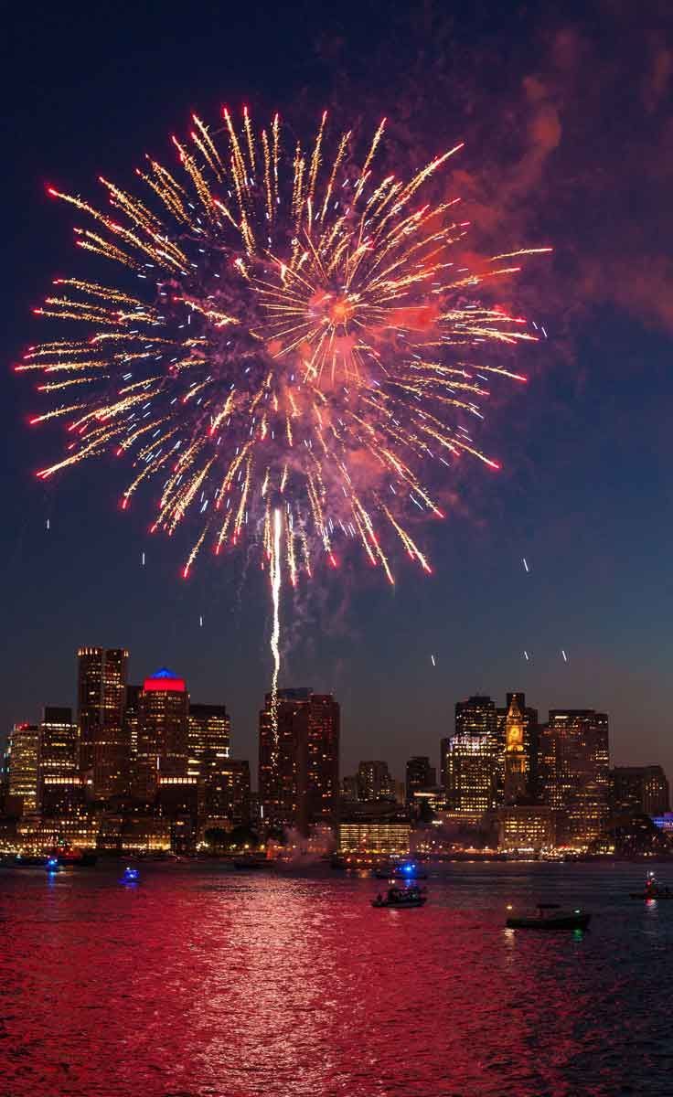 fireworks are lit up the night sky over water and cityscape in the background