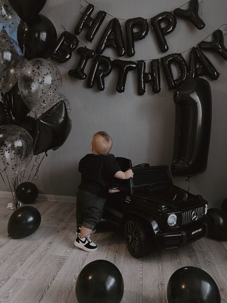 a young boy sitting on top of a black car in front of balloons and streamers