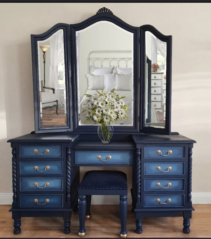 a blue dresser with two mirrors and a flower vase on top of it in front of a white bed