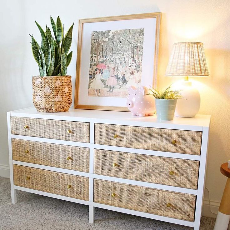 a white dresser with two plants on top and a framed photograph above it, next to a lamp