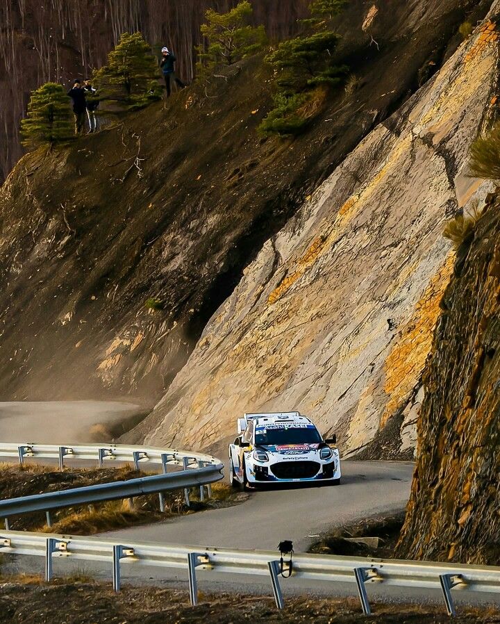 a white car driving down a road next to a mountain side with people standing on top of it